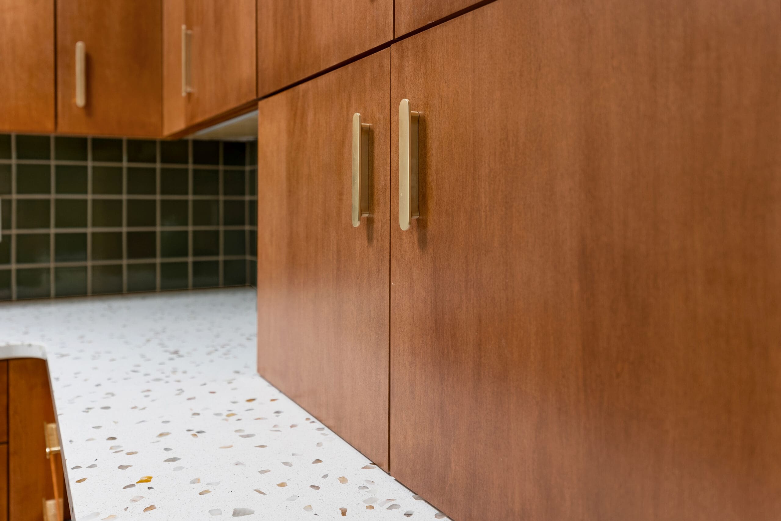 Close‑up of modern kitchen remodel with wood cabinetry, brass hardware, terrazzo countertop, and dark green tile backsplash.