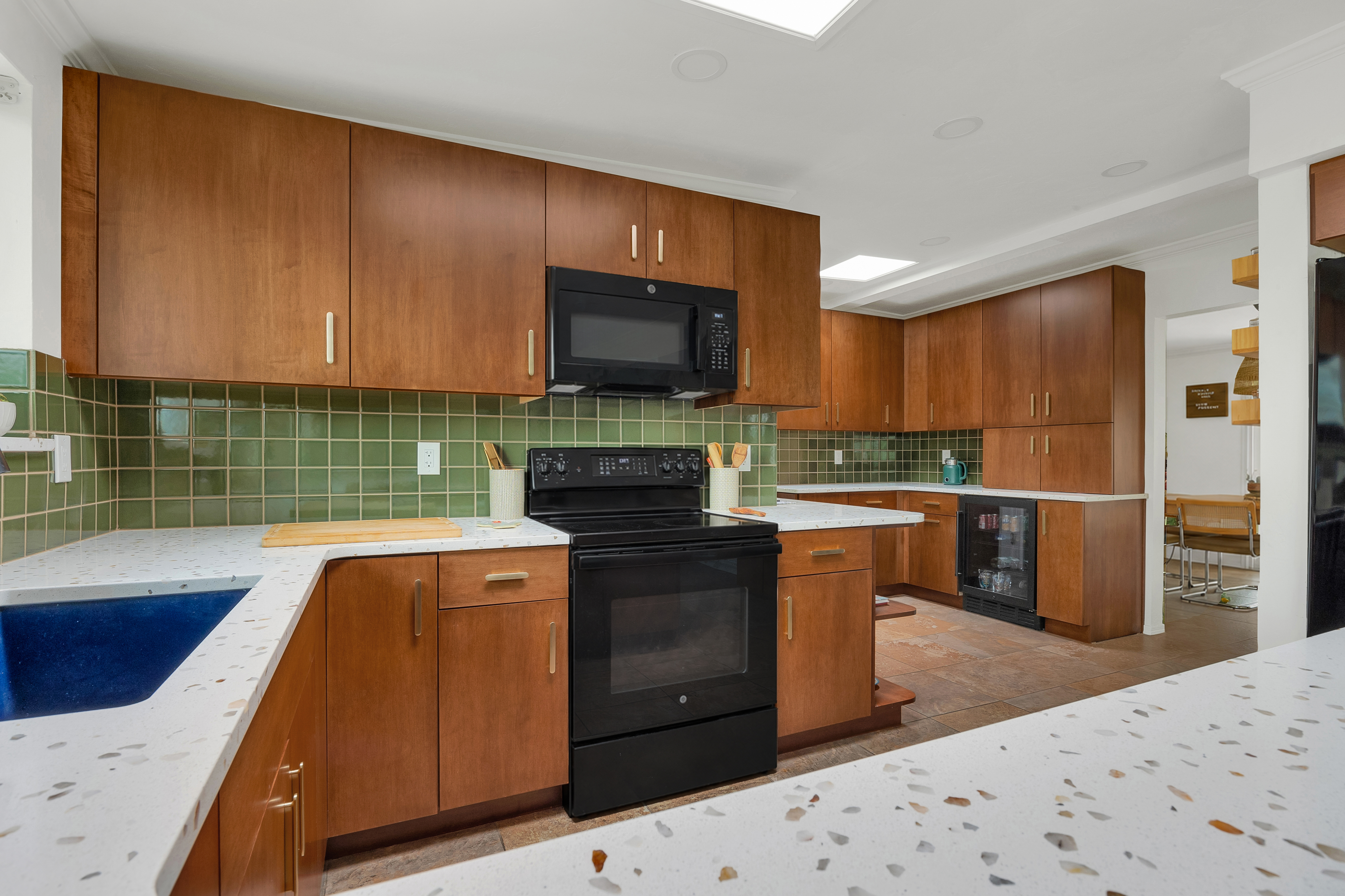 Modern kitchen remodel with wood cabinetry, terrazzo‑style countertops, green tile backsplash, and black appliances.