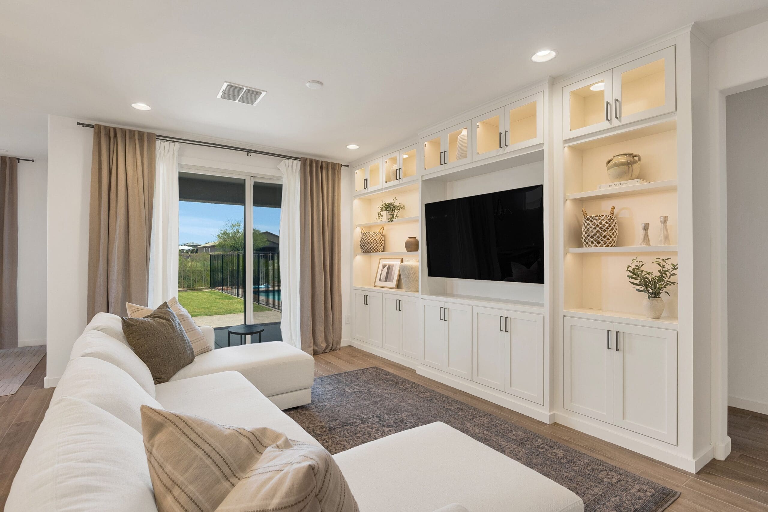 Modern living room with a custom white built‑in entertainment unit featuring illuminated shelves, centered TV, sectional sofa, sliding glass doors, and a view of the outdoor pool area.