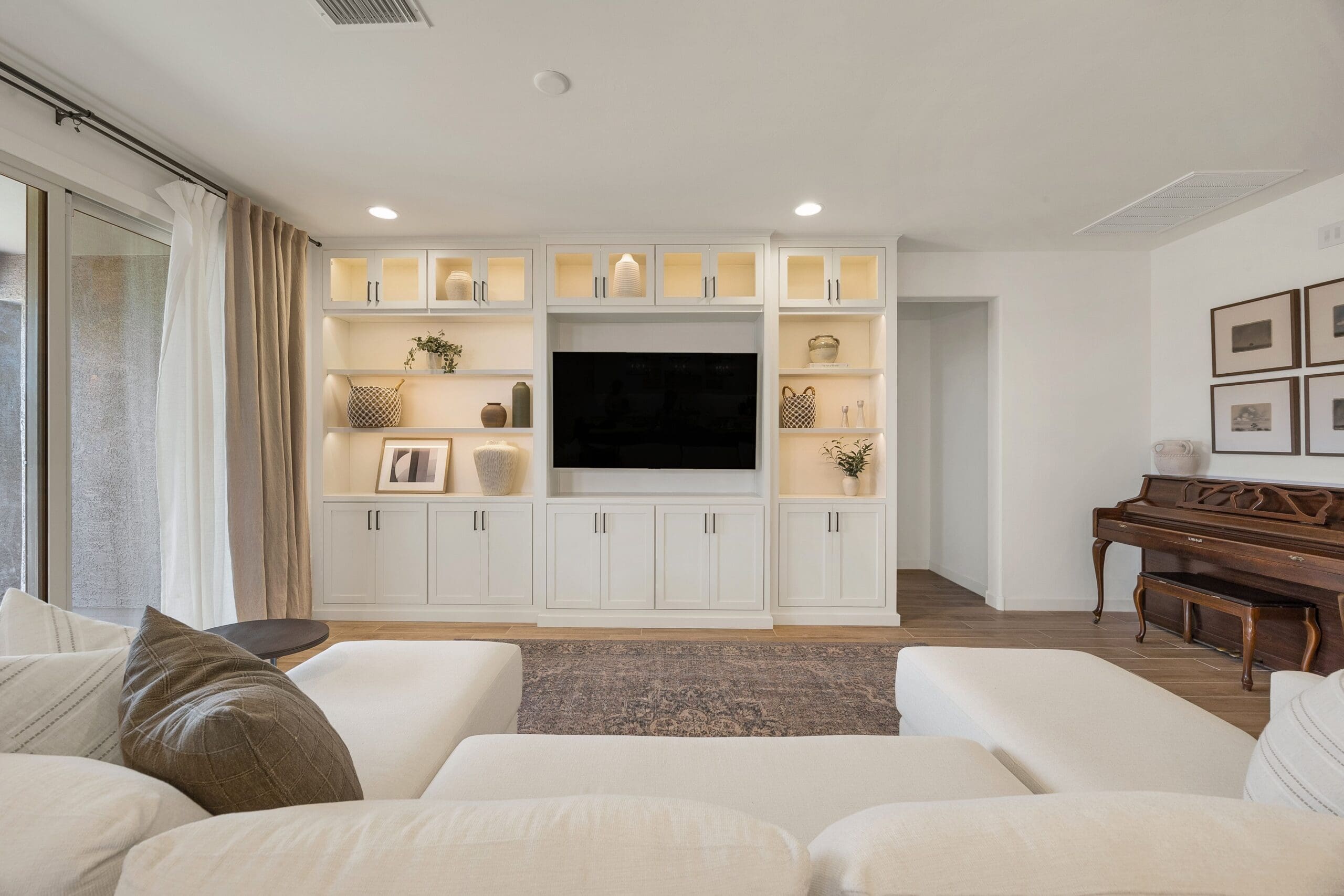 Modern white built‑in entertainment wall with Bertch cabinetry, integrated shelf lighting, glass‑front upper cabinets, and a centered TV surrounded by decor in a Tucson living room.