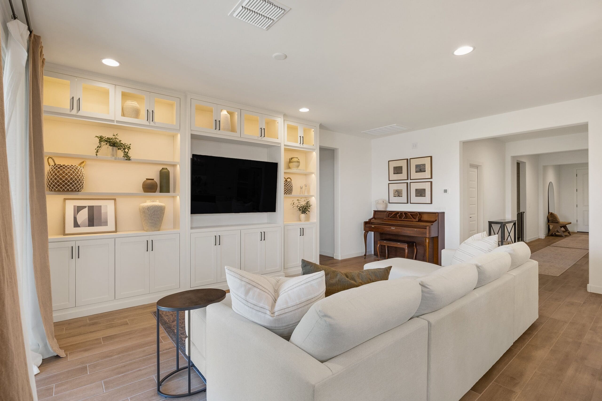 Modern living room with a custom white built‑in entertainment unit featuring illuminated shelves, centered TV, styled décor, sectional sofa, and adjacent piano.