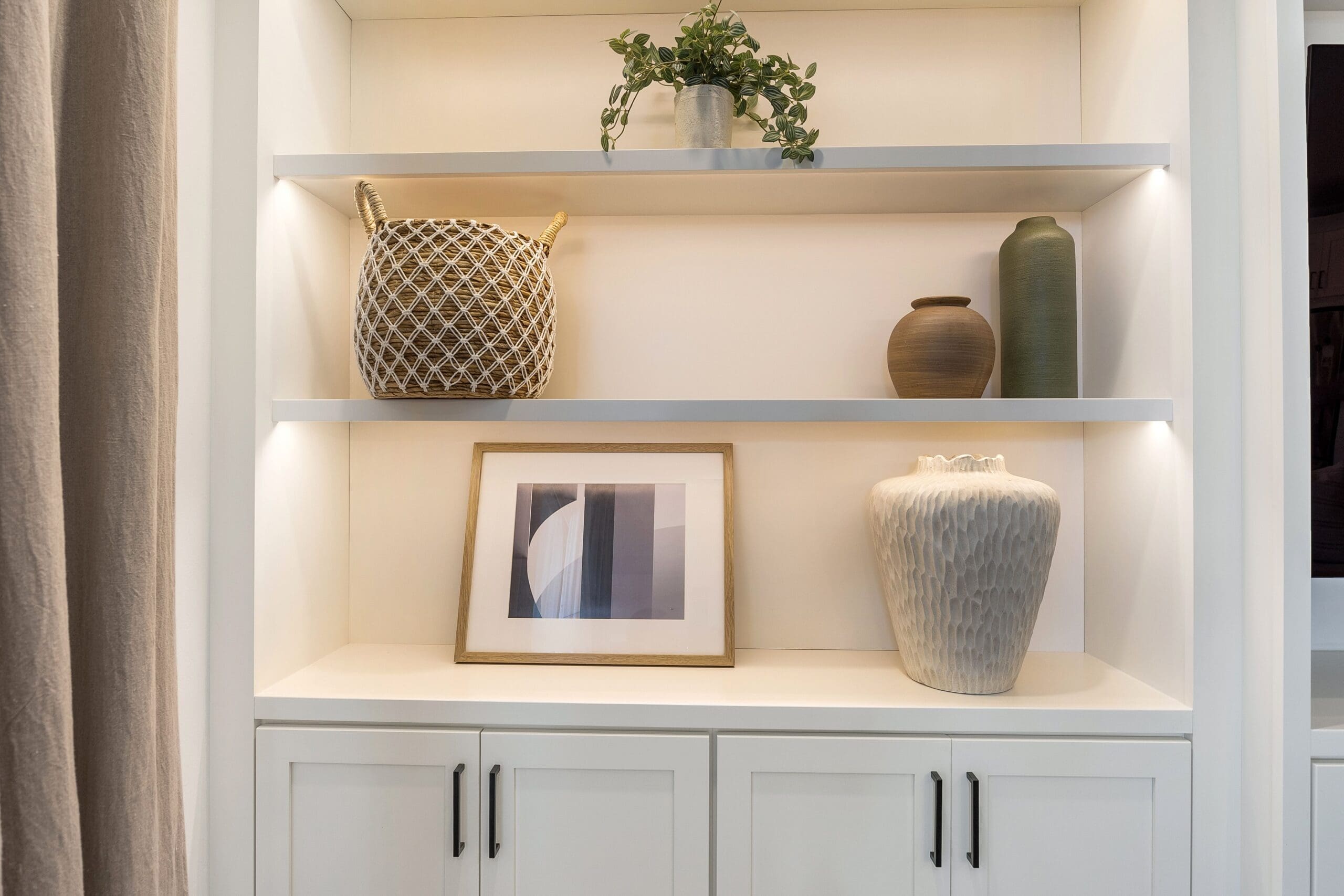 White built‑in shelving with custom cabinetry, decorative baskets, ceramic vases, framed artwork, and a potted plant, designed by Uncharted Design.