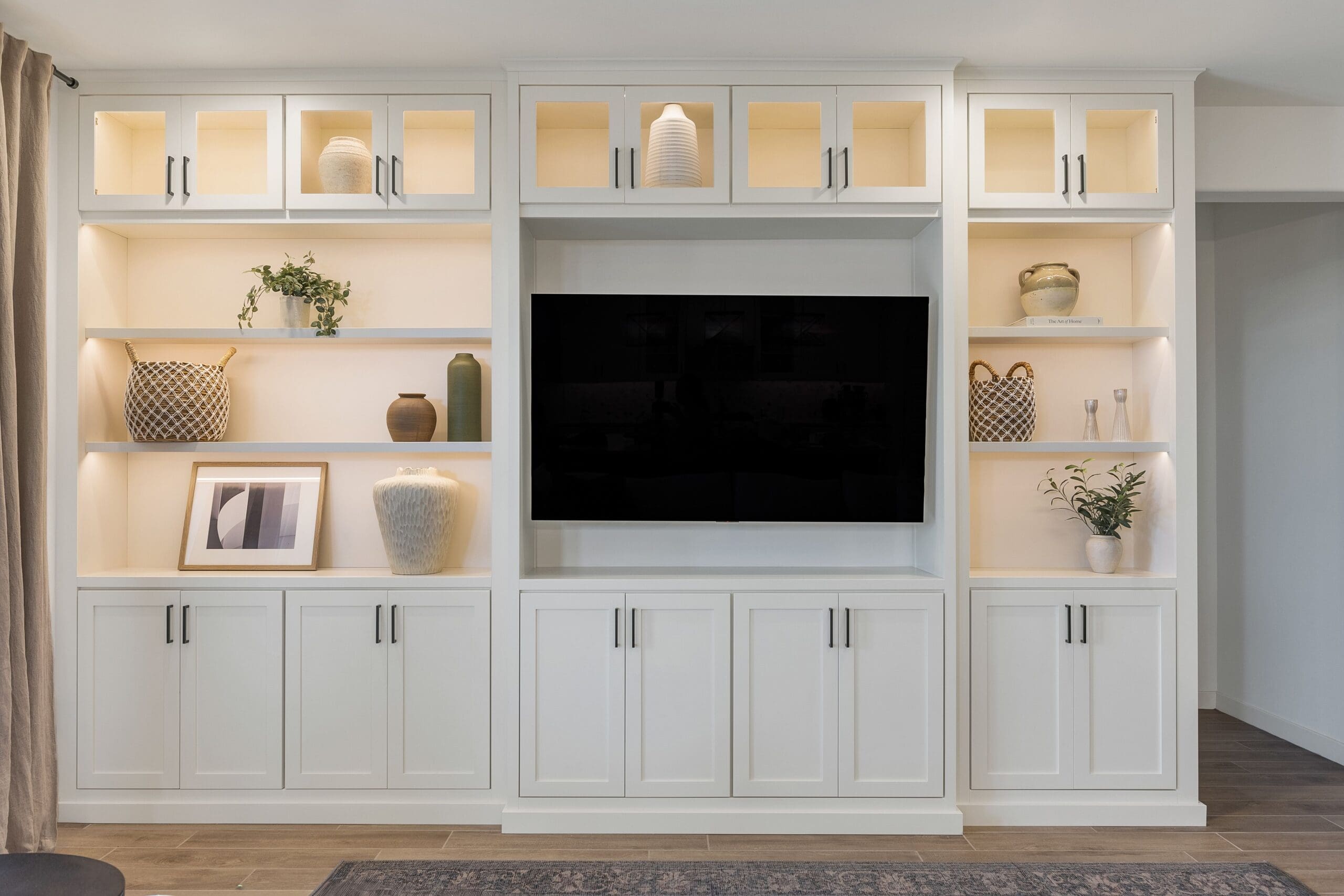 Modern living room with a custom white built‑in entertainment unit featuring illuminated shelves, centered TV, lower storage cabinets, sectional sofa, piano, and sliding glass doors.